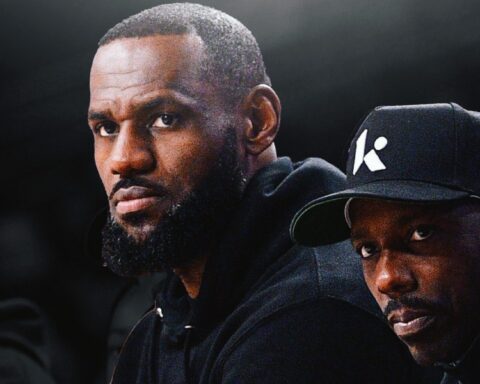 Two men in dark clothing pose closely against a dark background, one with a beard and the other wearing a cap with a white logo on the side.
