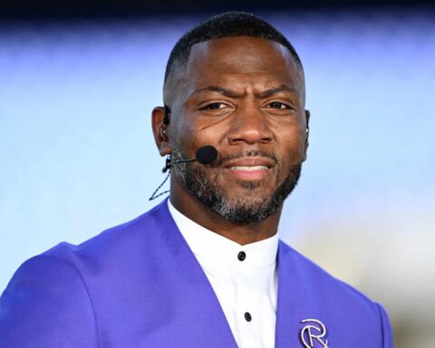 Portrait of a Black man wearing a purple suit and white shirt with a headset microphone, speaking at an event.