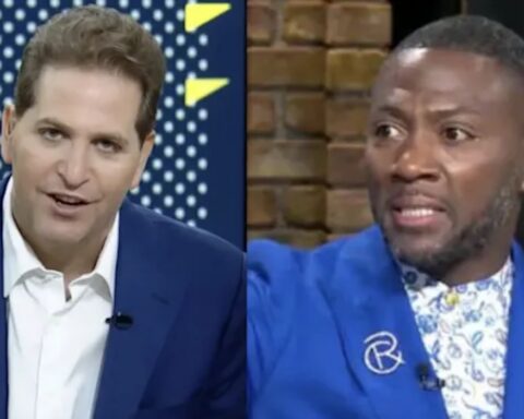 Split-screen of two men in a TV studio: left host in a blue suit speaks, right guest in a bright blue blazer gestures amid a brick background