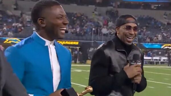 Two men on a football field during a broadcast, one in a bright blue suit and the other in a black jacket with a cap, both smiling.