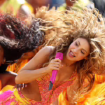 Front-center performer singing into a pink microphone while dancing in a vibrant orange-yellow festival scene with other dancers around her.