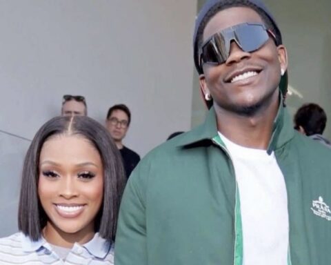 Smiling woman with a straight bob haircut posing beside a man in a green jacket and oversized sunglasses indoors, with people in the background.