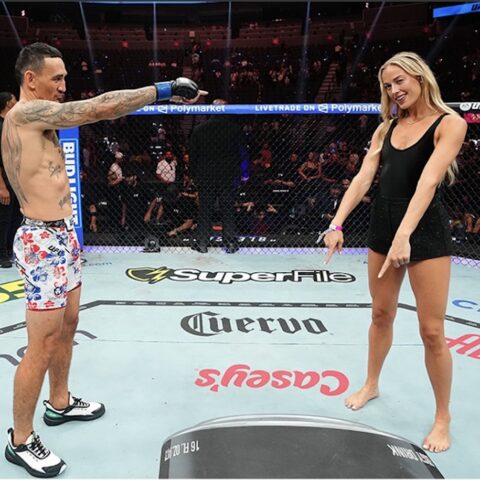 Two people stand in a UFC octagon facing each other; man points toward a woman in a black dress as spectators watch.
