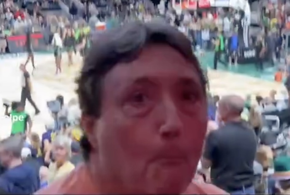 Close-up of a man with a flushed face in the foreground at a crowded sports arena, spectators behind him on bleachers.