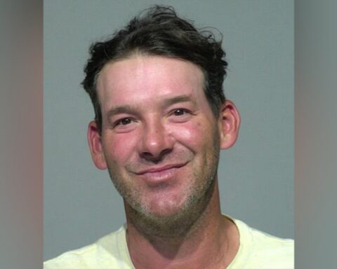 Mugshot-style portrait: man with short dark hair, light stubble, wearing a pale shirt against a gray backdrop, smiling slightly