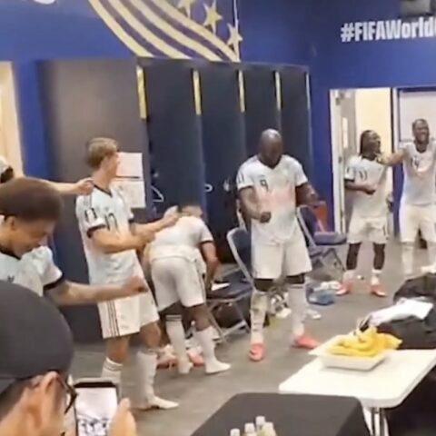 Soccer players in white uniforms celebrate in a locker room, several standing with arms raised and clapping near blue walls with #FIFA World Cup.