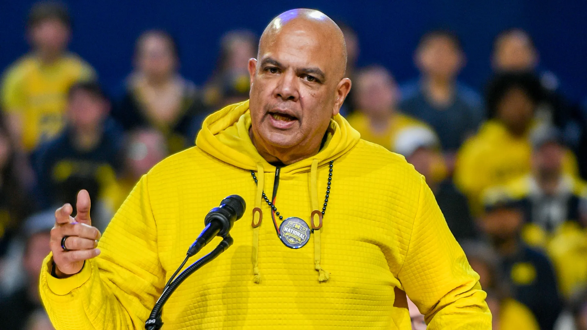 Man in a bright yellow hoodie speaks into a microphone at a crowded event, with blurred yellow-clad fans behind him.