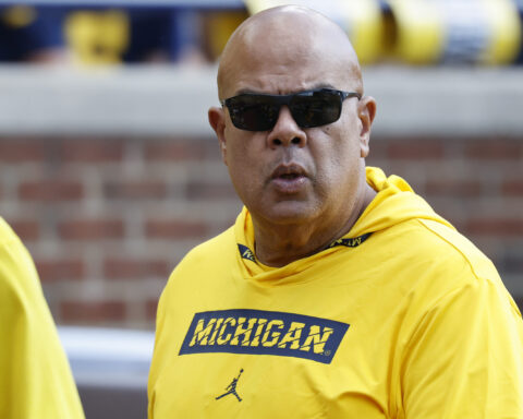 Man in a bright yellow Michigan shirt and dark sunglasses, speaking or mid-sentence outdoors.