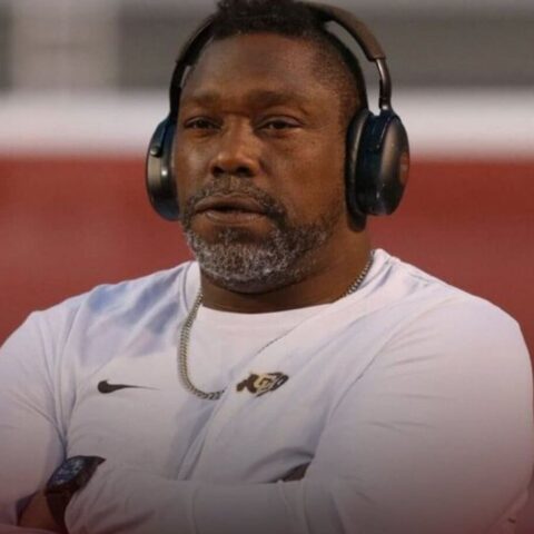 Man wearing large over-ear headphones and a white Nike shirt, arms crossed, in a stadium setting.