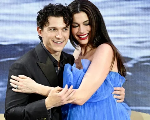 A smiling couple embraces on stage, the man in a black suit and the woman in a blue dress, backdrop of ocean scenery behind them.