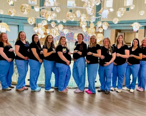 Group of pregnant women posing in a hospital lobby, wearing black tops and blue pants with hands on bellies.