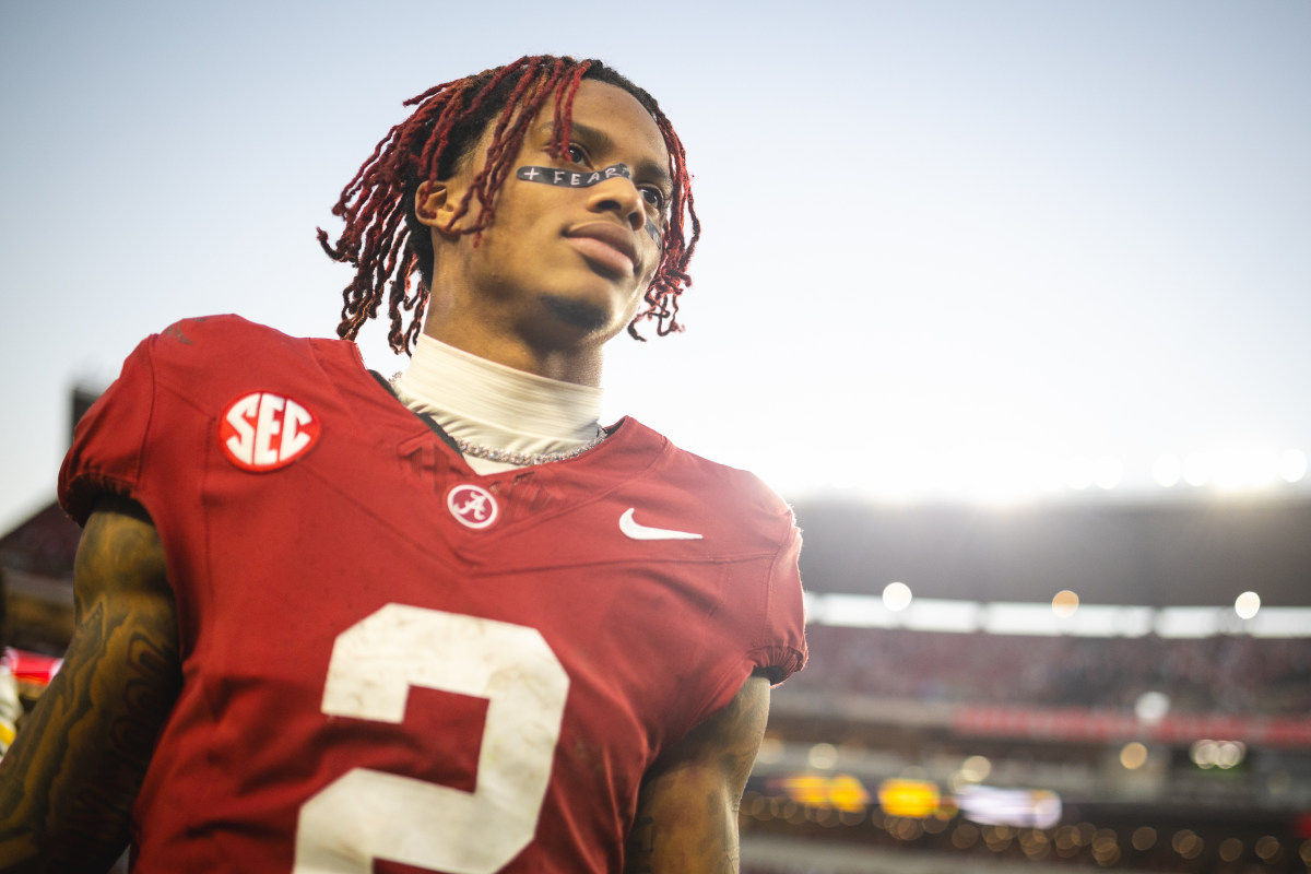 Young football player in a red jersey with SEC patch, standing on a stadium field under bright lights, looking upward with red dreadlocks and a face strip that reads '+ FEAR'.