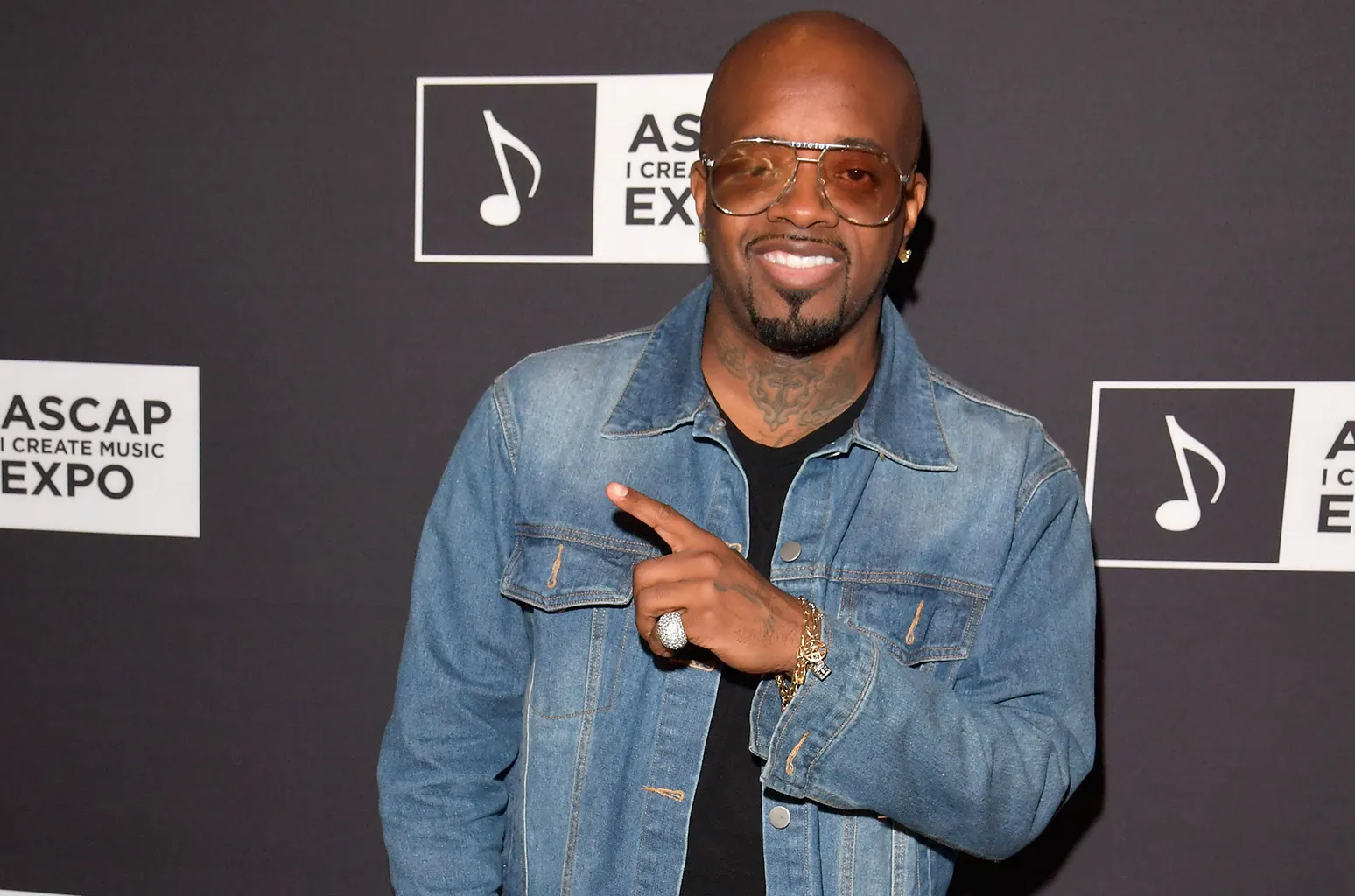Man wearing a denim jacket and sunglasses, pointing with his right hand in front of a music expo backdrop.