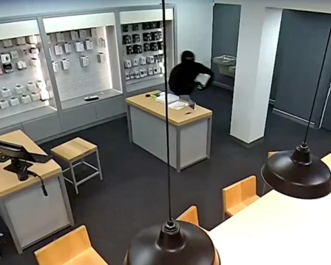 Security camera view of an electronics store: wall display with phone cases, a checkout counter with papers, and a masked shopper in dark clothing browsing a product shelf in the back corner.