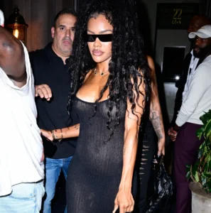 Woman with long curly hair in a tight black dress and sunglasses amid a crowded nightlife scene, carrying a small bag.