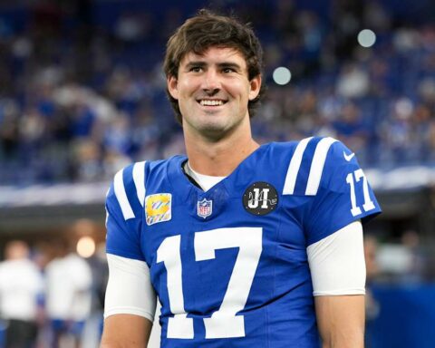 Football player in a blue jersey #17 smiling on the field with a blurred stadium crowd behind him.