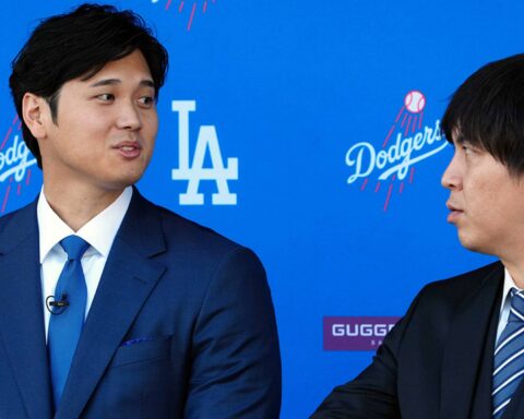 Two men in dark suits converse on a blue Dodgers backdrop, with one wearing a blue tie and the other a striped tie.