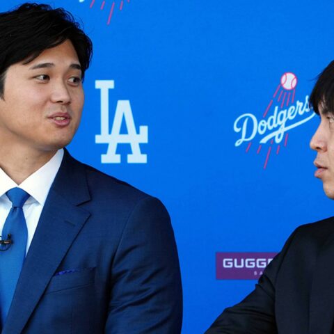 Two men in dark suits converse on a blue Dodgers backdrop, with one wearing a blue tie and the other a striped tie.