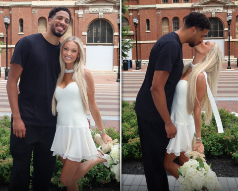 Couple posing outside a red brick market building; man in black, woman in a white dress holding white flowers.