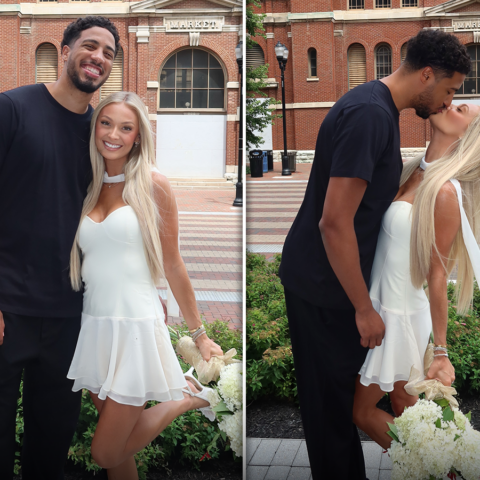 Couple posing outside a red brick market building; man in black, woman in a white dress holding white flowers.