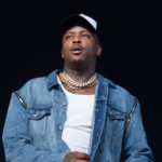 Man on stage wearing a white cap, heavy gold chains, and a blue denim jacket, looking upward against a dark background.