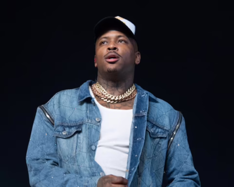 Man on stage wearing a white cap, heavy gold chains, and a blue denim jacket, looking upward against a dark background.