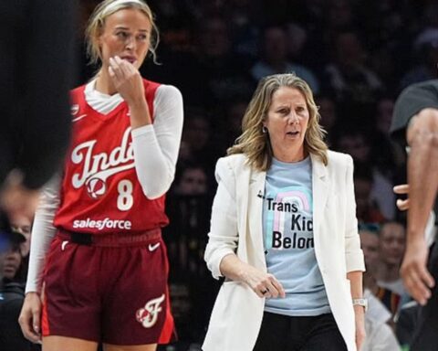Basketball coach in a white blazer and light blue 'Trans Kids Belong' shirt walking on the court beside a red Indiana jersey player.