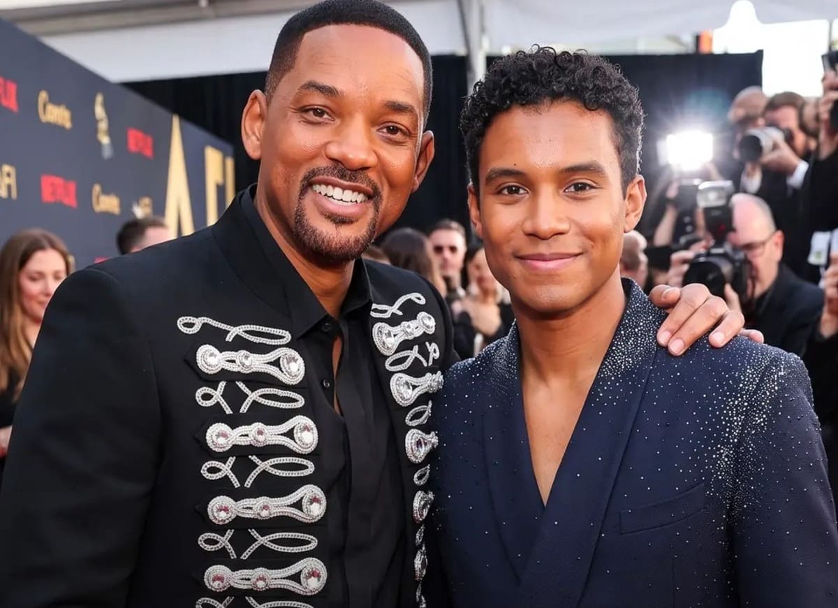 Two men on a red carpet posing for photos; one wears a black jacket with silver braiding, the other in a dark blue sequined suit, both smiling