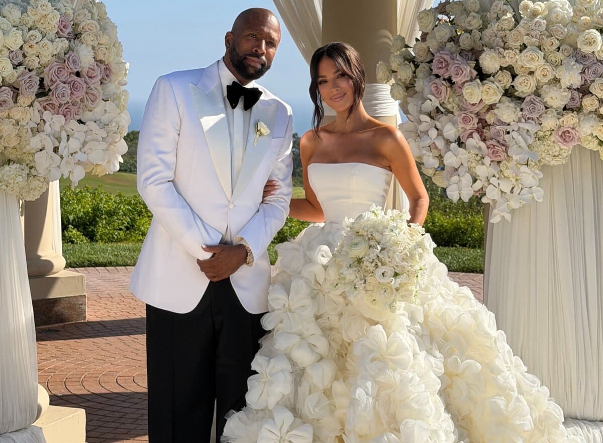 Bride in a strapless white gown with a voluminous floral train and groom in a white tuxedo jacket stand arm-in-arm under a cream and rose arch at an outdoor wedding ceremony.