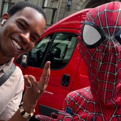 Man smiles for a selfie with someone dressed as Spider-Man beside a red van in an urban street.