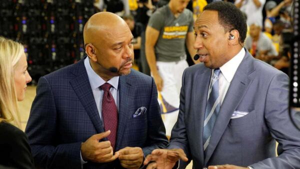 Two men in suits with wireless headsets talk on a basketball court, a woman partially visible on the left.