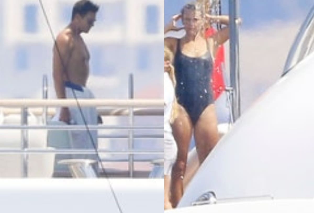 Man shirtless in light shorts standing on a yacht deck beside a woman in a black one-piece swimsuit by the railing on a sunny day.