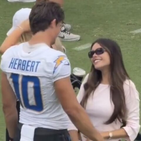 Football player Herbert #10 in a white jersey shakes hands with a smiling woman in a pink blouse on a football field.