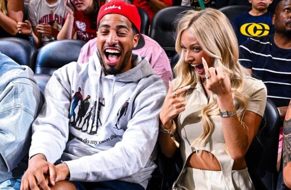 Two excited spectators: a man in a gray hoodie and red cap laughing, seated beside a blonde woman in beige, who is flashing a playful middle-finger gesture at a live event.