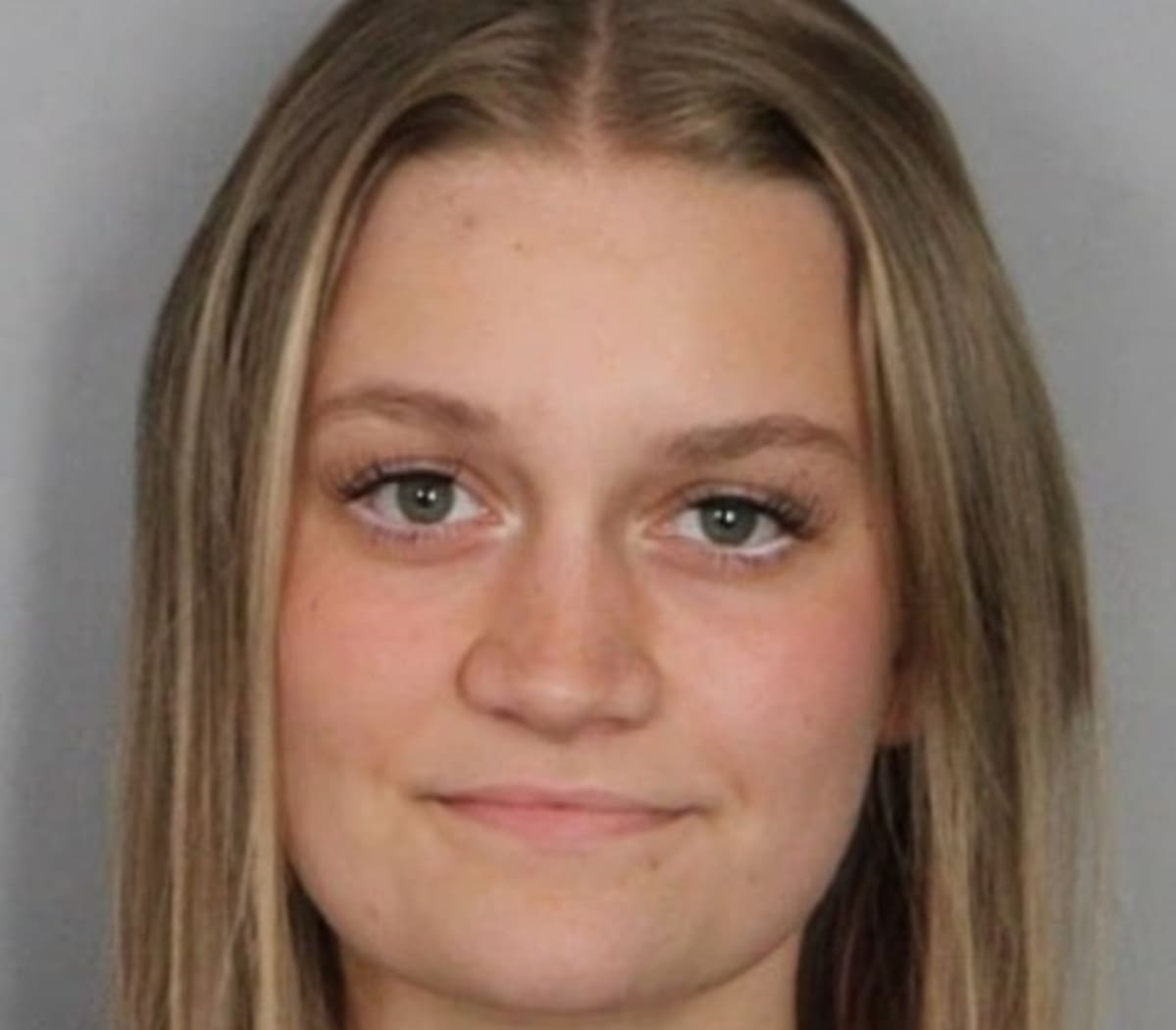Close-up portrait of a young woman with straight light brown hair and green eyes against a gray background, neutral expression.