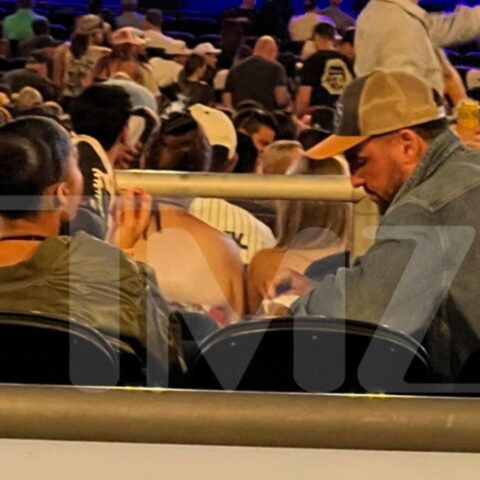 Two men seated in the foreground of a crowded stadium; one wears a beige cap and denim jacket. Behind them is a large audience under blue/purple lighting.
