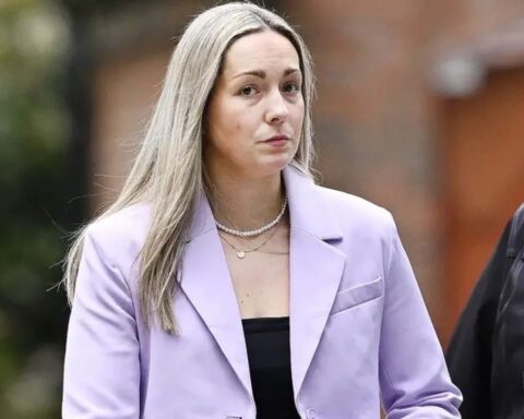 Woman with long blonde hair wearing a lavender blazer and pearl necklace, standing outdoors with a neutral expression.
