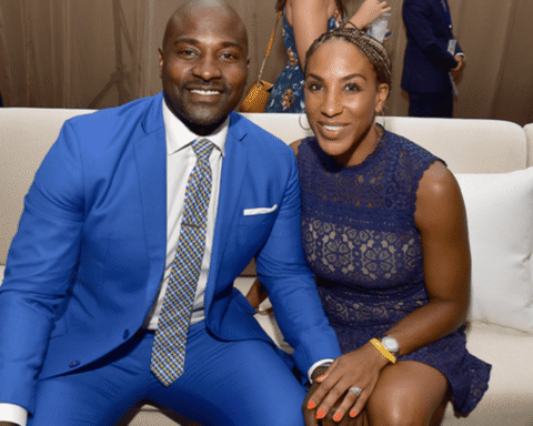 Smiling couple seated on a beige sofa at a formal event; man wears a bright blue suit and patterned tie, woman wears a navy lace dress.