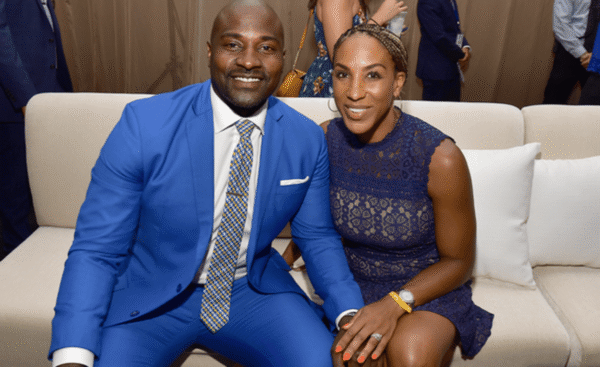 Smiling couple seated on a beige sofa at a formal event; man wears a bright blue suit and patterned tie, woman wears a navy lace dress.