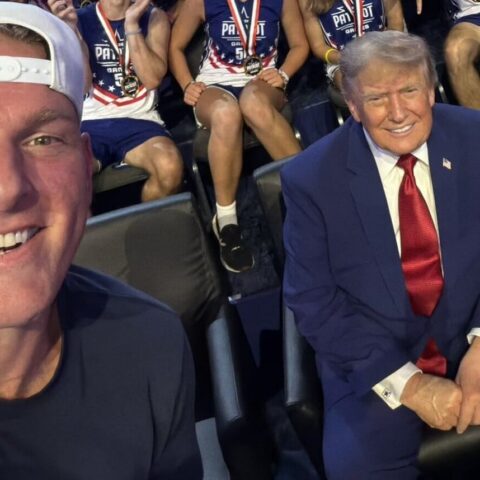 Close-up selfie of a smiling man wearing a white baseball cap, with a suited man in a navy suit and red tie seated beside him; patriotic athletes in the background.