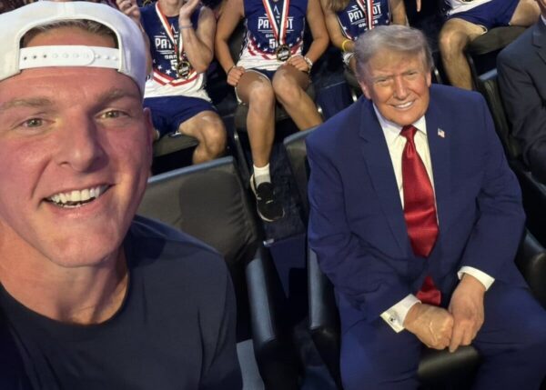 Close-up selfie of a smiling man wearing a white baseball cap, with a suited man in a navy suit and red tie seated beside him; patriotic athletes in the background.