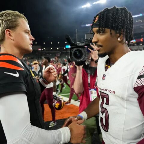 Two football players shake hands on the field after a game, facing each other, with a camera crew in the background at night.