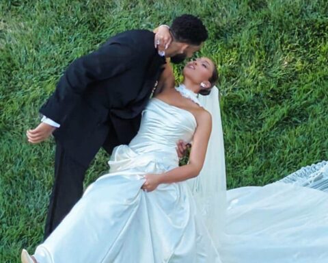 Bride in a white wedding dress leans back as a groom in a black suit kisses her on a green lawn.