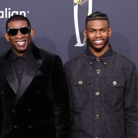 Two men pose on a red carpet at a formal event; left man wears a black velvet double-breasted suit and sunglasses, right man wears a dark buttoned jacket.