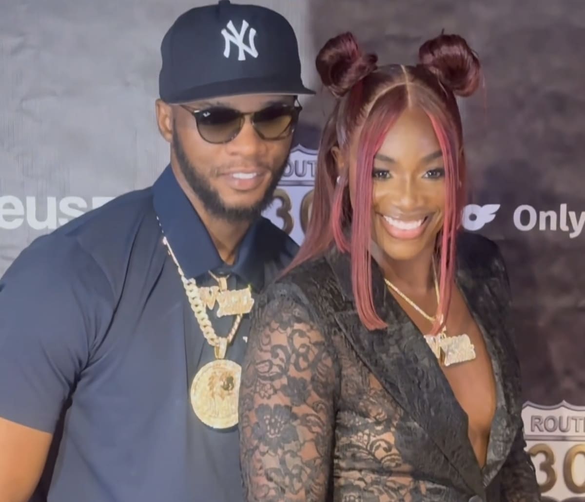 A smiling couple posing together at a red-carpet event; man in a NY cap and sunglasses with gold chains, woman in a black lace outfit with pink hair buns.