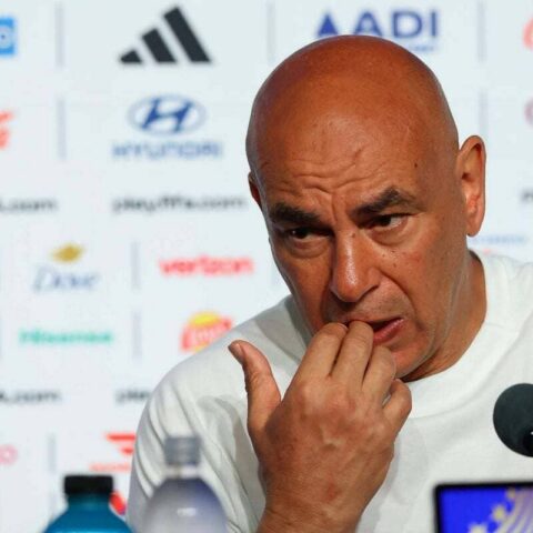 Bald man in a white shirt at a press conference, touching his lips, with a microphone and sponsor logos in the backdrop.