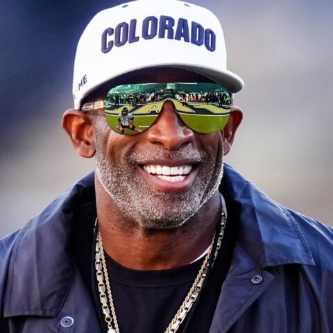 Smiling African American man wearing a white Colorado cap, dark jacket, and gold chains, with mirrored sunglasses reflecting a football field and players.
