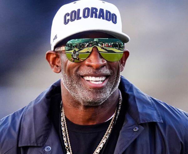 Smiling African American man wearing a white Colorado cap, dark jacket, and gold chains, with mirrored sunglasses reflecting a football field and players.