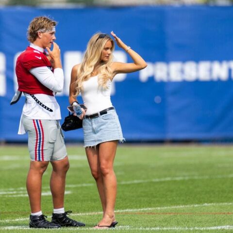 Football player in red and white gear stands with a casually dressed woman on a grassy field, helmet resting on the turf nearby.