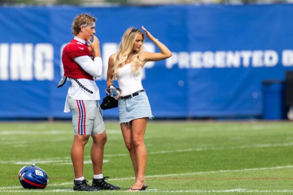 Football player in red and white gear stands with a casually dressed woman on a grassy field, helmet resting on the turf nearby.